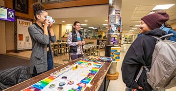 A Saint Michael's College student stands at a table in the Alliot Student Center and takes a photo of two students. 