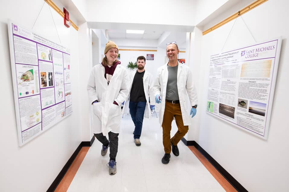 Professor Mark Lubkowitz wears a white lab coat abd walks down a hall way with two students also wearing white lab coats.
