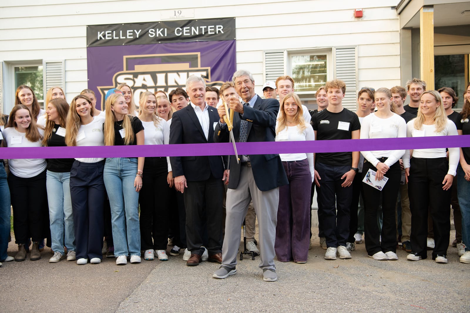 Tom Kelley ’69, the namesake of the new Kelley Ski Center, raises the giant pair of scissors after cutting the ribbon during the grand opening of the center on Sept. 20, 2024. President Richard Plumb, left, and members of the Alpine and Nordic skiing teams applaud.